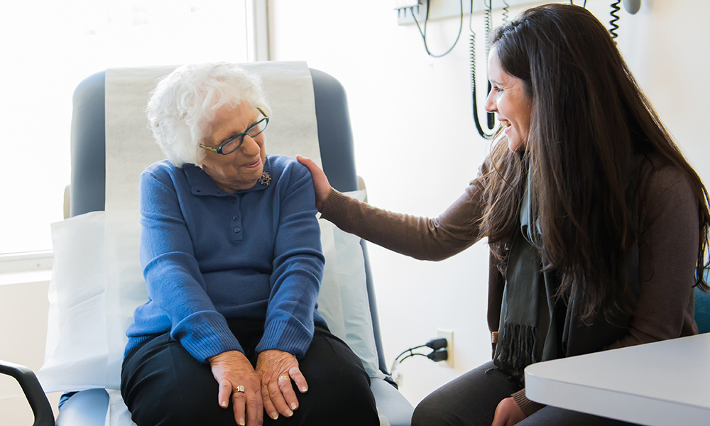 A smiling older woman sits in an exam chair while a younger woman sits beside her, hand on her shoulder.